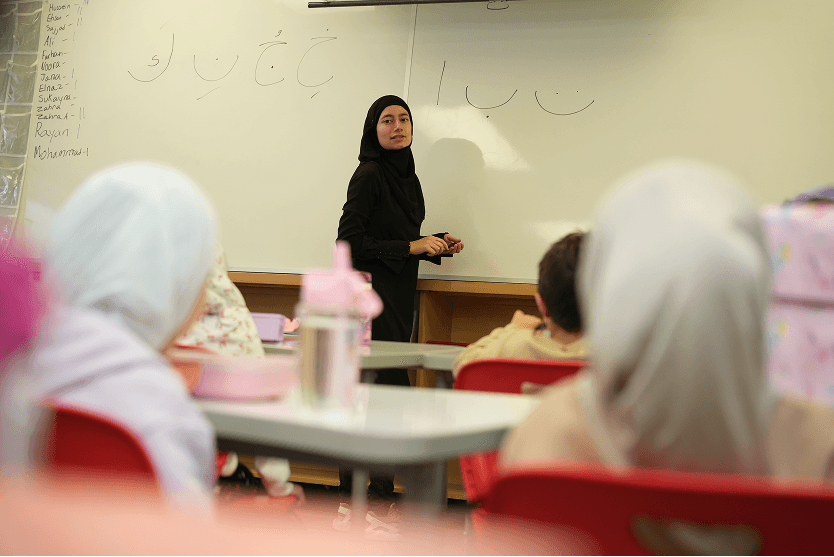 Female teacher in a black hijab teaching Arabic script to children in a classroom.