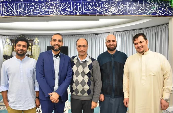 Five men standing side by side indoors with smiling expressions, traditional Arabic calligraphy displayed above them.