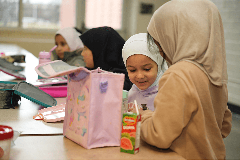 Four young girls wearing hijabs sitting at a table with lunchboxes and a juice box, sharing a moment together.