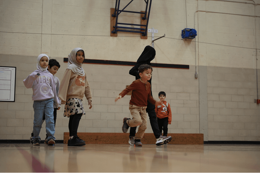 Five children, including two wearing hijabs, playing or running in an indoor gymnasium with a beige brick wall background.