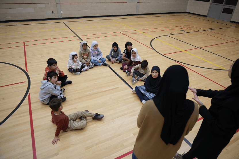 A group of children sitting on the floor in a gymnasium, listening to two women standing and talking to them.