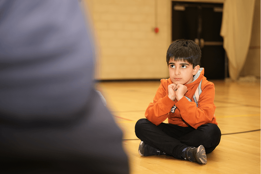 Young boy in orange hoodie sitting cross-legged on a gym floor, looking attentively at someone off-frame.